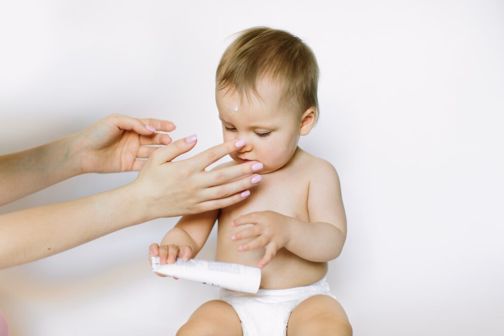 Mother hands applying cream on baby after bathing on white background. The baby holds the tube with